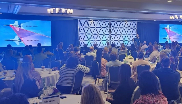 Eleven Eleven AV large-scale conference deployment in a San Diego ballroom. Synchronized LED screens display an intricate graphic analyzing "Blind Spots" using jet fighter imagery, while an executive speaks. This setup supports a nationwide live stream with triple-layer redundancy for a Fortune 500 client.