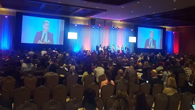 The packed Los Angeles conference center for a national franchise intensive, with 1,200 attendees seated. The stage features a live executive panel magnified on twin high-fidelity projection screens, demonstrating Eleven Eleven AV's visual parity engineering and master-led multi-camera switching for a fault-tolerant 14-state virtual broadcast.