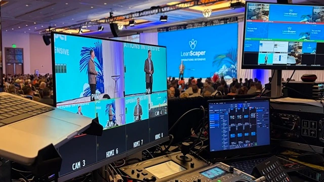 A high-end live event production booth featuring a digital video switcher, multi-view camera monitors, and technical control laptops during a corporate leadership summit.